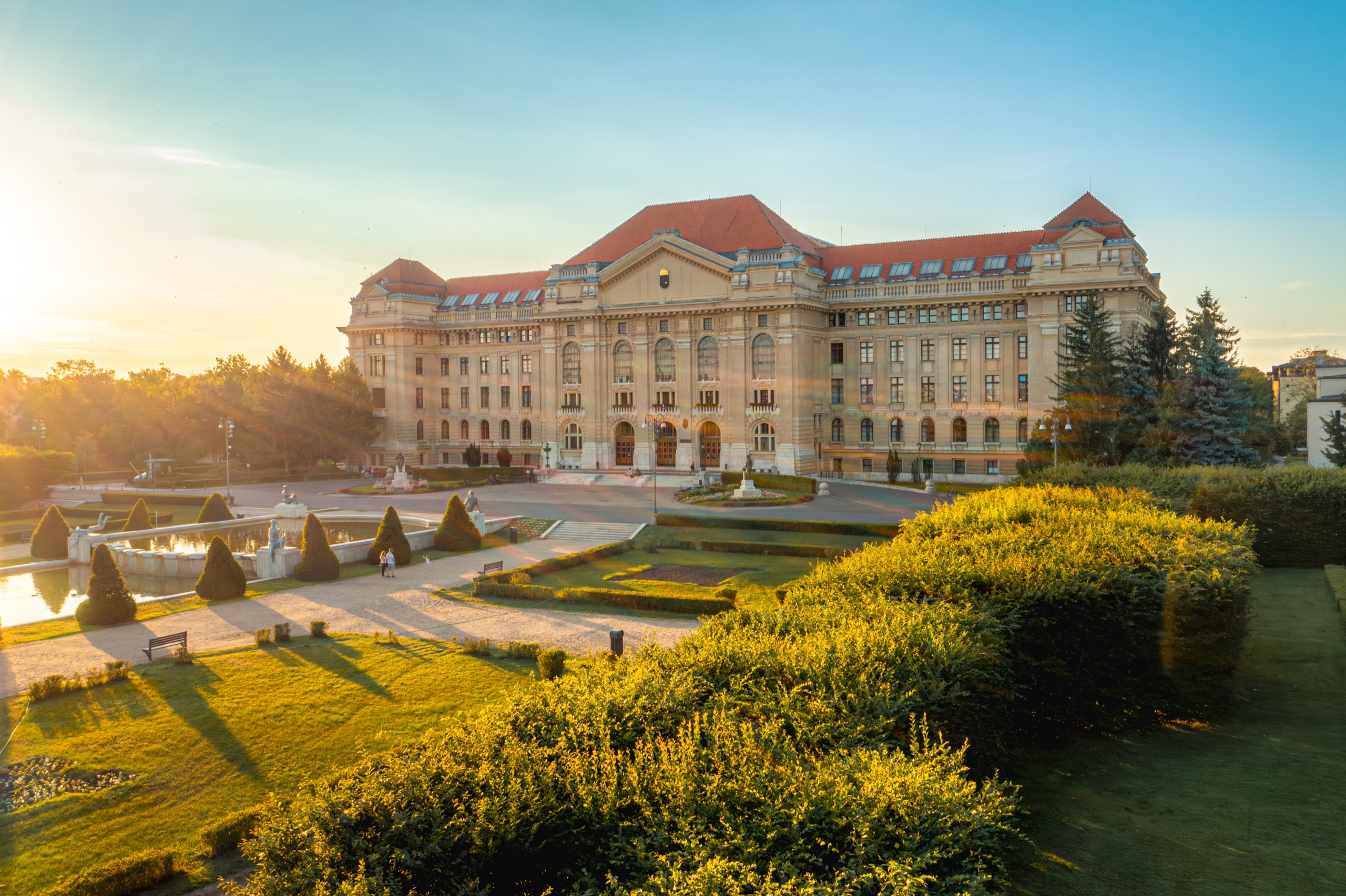 A Hungarian medical university main building bathed in warm golden-hour light, clouds drifting and the fountains shimmering.